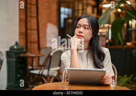 Serious Asian woman in white dress looking away using tablet while sitting at table in cafe Stock Photo