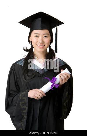Smiling Chinese woman wearing graduation gown and cap holding diploma while standing against white background in studio Stock Photo