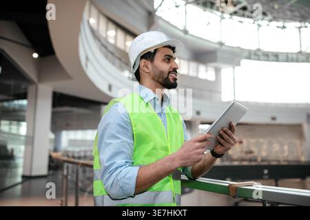 Middle Eastern male engineer wearing hard hat and vest looking away and holding tablet while standing beside terrace in office Stock Photo