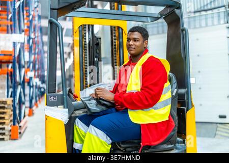 Black male worker wearing safety vest looking away driving forklift in distribution warehouse Stock Photo