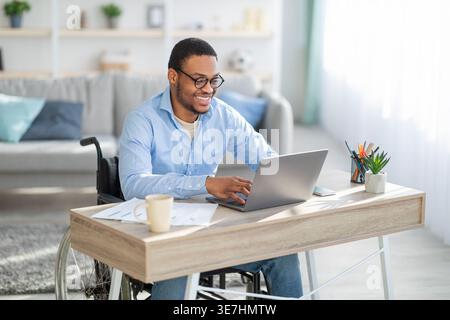 Smiling Black disabled man wearing eyeglasses working on laptop while sitting at table in home office Stock Photo
