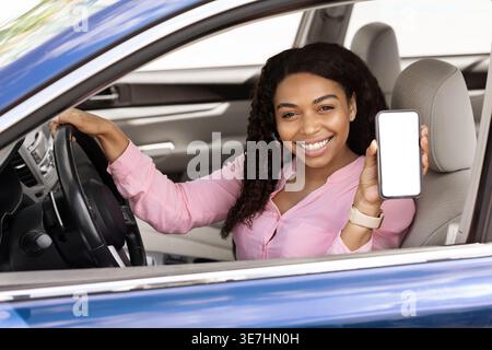 Smiling Black woman wearing pink shirt showing smartphone while sitting on driver seat holding steering wheel in car Stock Photo
