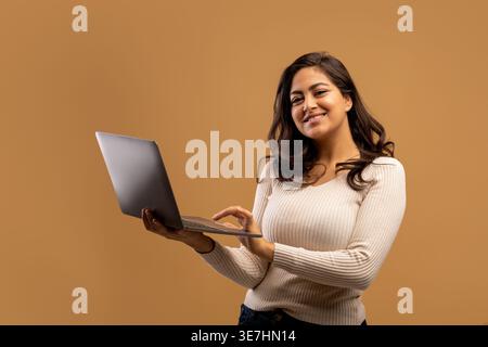 Smiling Middle Eastern woman wearing holding laptop standing against brown background in studio Stock Photo
