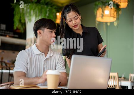 Thai woman standing and sharing tablet with male colleague discussing project details while sitting at table with laptop in cafe Stock Photo