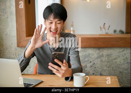 Smiling Asian man waving hand having video call on smartphone sitting at table with laptop and coffee cup at home Stock Photo