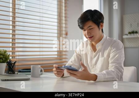 Smiling Asian man holding card and smartphone using mobile banking application sitting at table at home during daytime Stock Photo