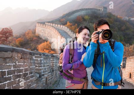 Smiling Chinese couple taking picture using digital camera looking away while standing on Great Wall during daytime Stock Photo