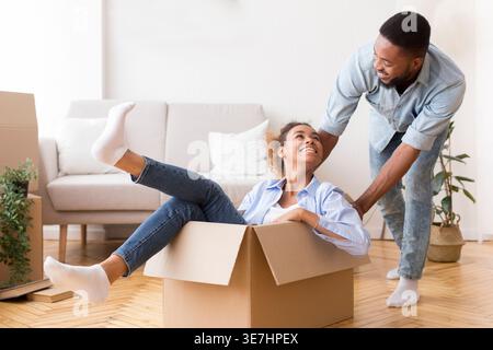Cheerful Black man having fun pushing wife sitting in moving boxes in new home Stock Photo