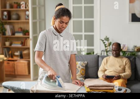 Black young female social service worker wearing uniform ironing clothes standing near middle-aged Black man sitting on sofa and using smartphone i... Stock Photo