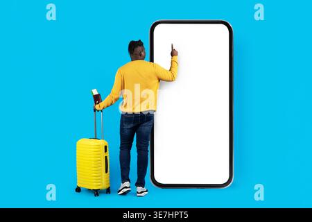 Black man with luggage holding passport and boarding pass using big smartphone standing in front of blue background in studio Stock Photo