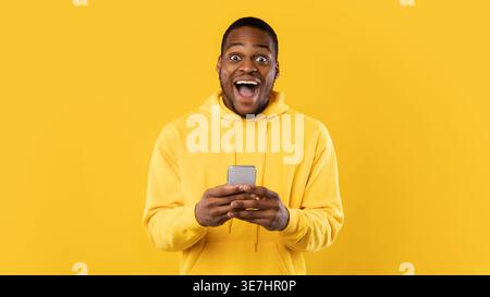 Excited bearded Black man wearing hoodie holding smartphone with both hands while standing with mouth opened in front of yellow background in studio Stock Photo