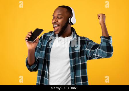 Black man wearing headphones singing using smartphone while standing against yellow background in studio Stock Photo