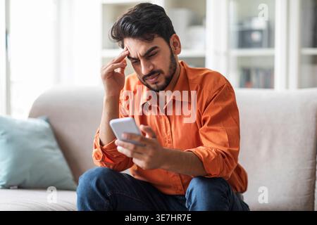 Stressed Middle Eastern man touching temple looking at smartphone in his hand sitting on couch at home Stock Photo
