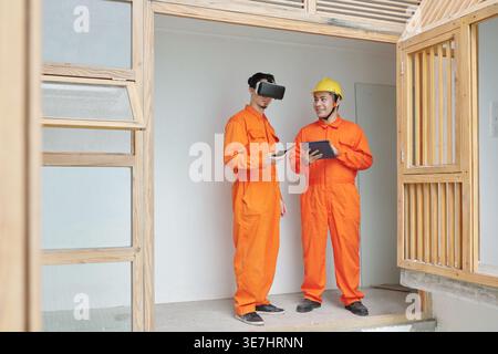 Mixed race male construction worker using virtual reality headset talking to Asian male construction worker holding tablet while standing together ... Stock Photo