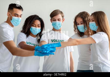 Smiling group of volunteers wearing face masks and gloves stacking hands together while standing against white wall in studio Stock Photo