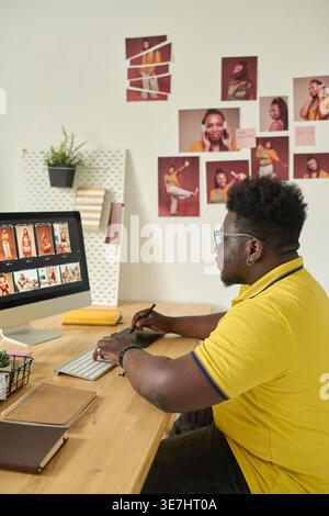 Black male photo editor wearing yellow polo shirt using graphic tablet for retouching photos on computer while sitting at table in office Stock Photo