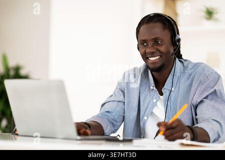 Smiling Black man wearing headset taking notes and using laptop while sitting at table in living room at home Stock Photo