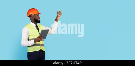 Black male engineer wearing hard hat and safety vest holding tablet looking away gesturing hand while standing against blue background in studio Stock Photo