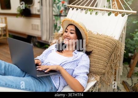 Asian woman wearing hat using laptop lying on hammock near motorhome during camping trip in forest Stock Photo