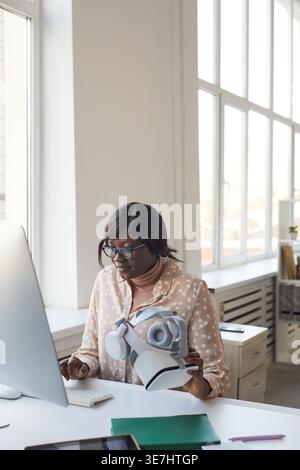 Black businesswoman wearing eyeglasses holding virtual reality headset working on computer while sitting at desk in office Stock Photo