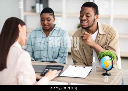 Confused Black couple looking at expensive family tour offered by caucasian female travel agent while sitting at table in tour agency office Stock Photo