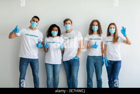 Smiling group of volunteers wearing face masks and gloves showing thumbs up sign while standing against white wall in studio Stock Photo