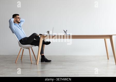 Relaxed Middle Eastern man in eyeglasses leaning back with hands behind head looking at laptop screen sitting at table in office during break Stock Photo
