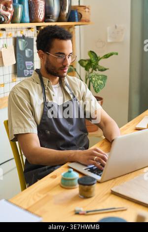 Black craftsman working on laptop sitting at table in front of porcelain tableware and sketches in workshop Stock Photo