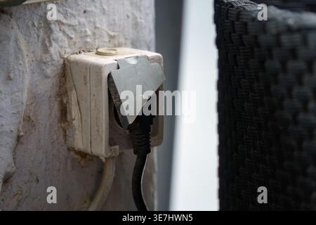 Old electrical outlet with a black plug and damaged cover mounted on a whitewashed wall. Stock Photo