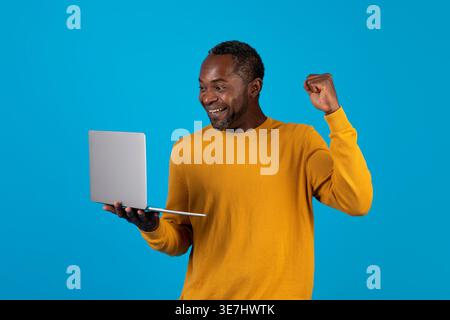 Cheerful Black man with closed fist using laptop celebrating success while standing against blue background in studio Stock Photo