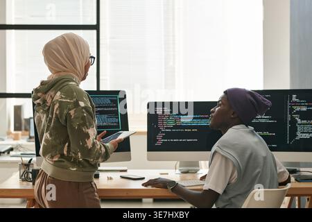 Black male and Mixed race Muslim female software developers discussing work near table with computer monitors in office Stock Photo