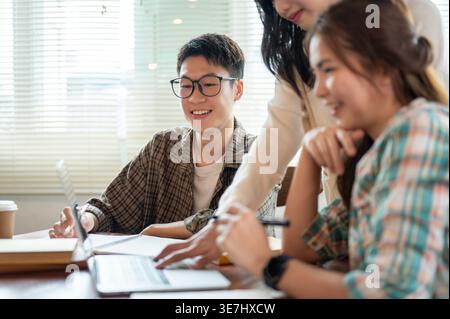 Smiling Asian male college student wearing eyeglasses sitting at table and working on school project with female classmates using laptop in cafe Stock Photo