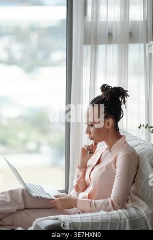 Black woman with locks looking at laptop screen thinking while sitting on sofa near glass window in her apLifestylement building Stock Photo