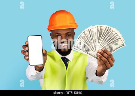 Black male engineer wearing hard hat and safety vest showing money and smartphone with blank screen while standing against blue background in studio Stock Photo