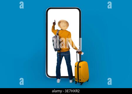 Black man with backpack wearing hat using big smartphone holding passport and tickets while standing with luggage against blue background in studio Stock Photo