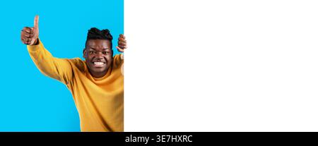 Cheerful Black man showing thumbs up hiding behind white blank advertisement board standing against blue background in studio Stock Photo