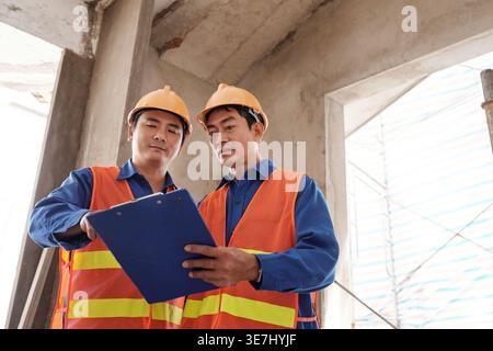 Asian male construction workers discussing over document in clipboard while standing together in construction site Stock Photo