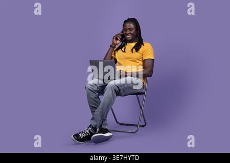 Smiling Black man with dreads wearing yellow shirt using laptop and having phone call while sitting on chair against purple background in studio Stock Photo