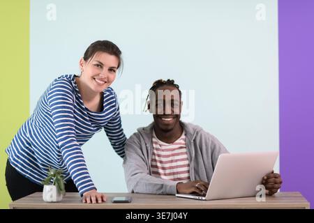 Smiling Black businessman with braids sitting at table with laptop near caucasian female colleague standing beside him against colorful wall in the... Stock Photo