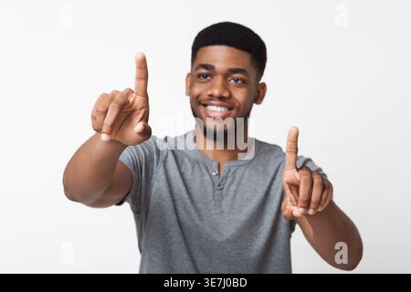 Smiling Black man wearing gray shirt touching invisible screen while standing against white background in studio Stock Photo