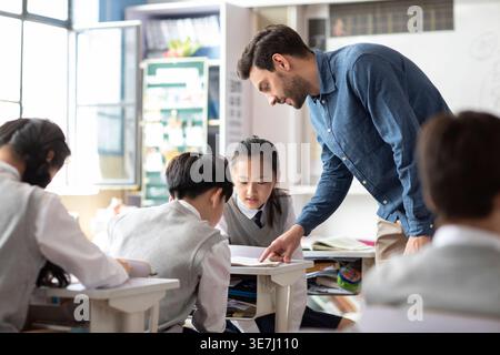 Diverse group of students doing school work while caucasian male teacher assisting them in classroom Stock Photo