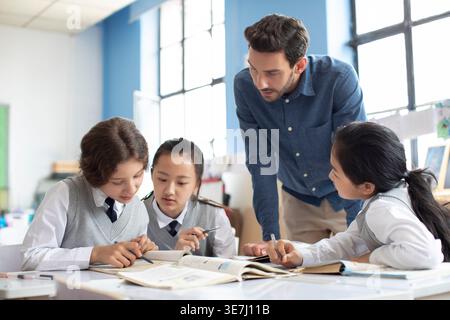 Diverse group of students doing school work with caucasian male teacher assisting them in classroom Stock Photo