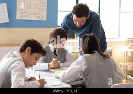 Diverse group of students doing school work while caucasian male teacher assisting them in classroom Stock Photo