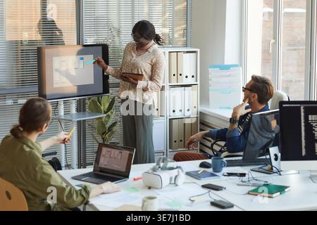 Black female programmer wearing eyeglasses holding tablet presenting in front with caucasian male colleagues while standing in office Stock Photo