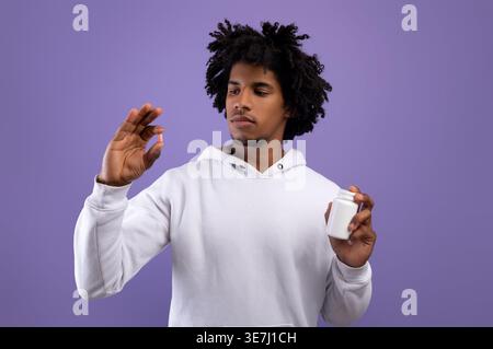 Black man with afro wearing white hoodie holding pill and medicine bottle while standing against purple wall in studio Stock Photo