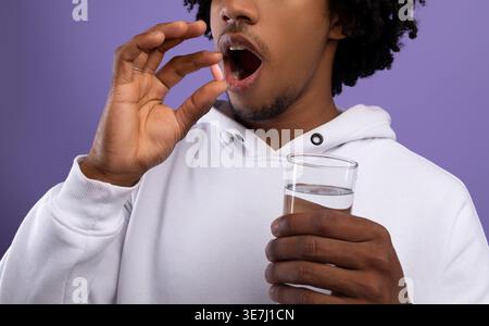 Black man with afro wearing white hoodie taking pill medicine holding glass of water while standing against purple wall in studio Stock Photo