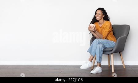 Middle Eastern woman with curly hair looking away holding mug while sitting on chair against white background in studio Stock Photo