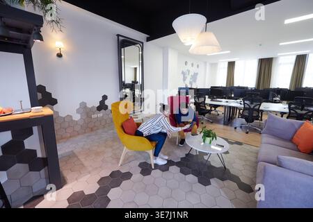 Middle Eastern male watching on tablet with caucasian female coworker sitting on sofa in office relaxation area during break Stock Photo