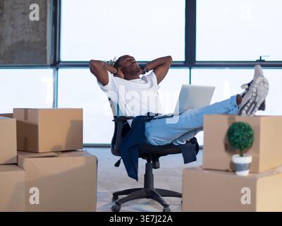 Black male architect with closed eyes relaxing with hands behind head and feet up on boxes while sitting in a new unfinished startup office Stock Photo