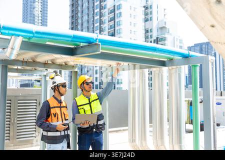 Asian male engineer wearing safety helmet pointing finger away holding laptop showing project details to Asian male coworker standing near pipeline... Stock Photo